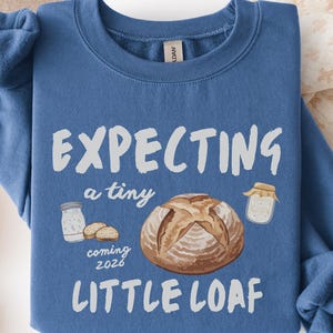 Puede incluir: Una sudadera azul con el texto "EXPECTING a tiny LITTLE LOAF" en blanco. El diseño incluye ilustraciones de una hogaza de pan, un tarro de harina y el año "2026". La sudadera tiene cuello redondo y mangas largas.