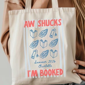 Puede incluir: Una bolsa de tela color crema con el texto en rojo AW SHUCKS e I'M BOOKED. Entre el texto hay ilustraciones de línea azul de libros abiertos, ostras y marcapáginas. El texto Summer 2026 y Charlotte está impreso en una tipografía azul.