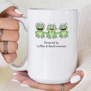 Könnte beinhalten: Weiße Keramik-Tasse mit drei Cartoon-Fröschen mit verschiedenen Gesichtsausdrücken. Auf der Tasse steht der Text "Powered by Coffee & Reinforcement". Die Tasse wird von einer Person gehalten, die einen weißen Pullover trägt.