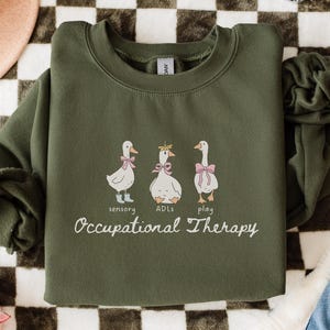 May include: An olive green sweatshirt featuring three cartoon geese with pink bows and the words "sensory", "ADLs", and "play" above the text "Occupational Therapy". The sweatshirt is folded on a checkered surface.