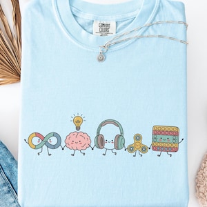 Puede incluir: Una camiseta azul claro con un gráfico de cinco personajes antropomórficos tomados de la mano: un símbolo de infinito, un cerebro con una bombilla, auriculares, un fidget spinner y un juguete pop-it cuadrado. La camiseta tiene una etiqueta Comfort Colors.