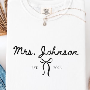 Puede incluir: Una camiseta blanca de cuello redondo con texto negro que dice Mrs. Johnson, un gráfico de lazo centrado debajo del nombre y el texto EST. 2026 a ambos lados del lazo. La camiseta tiene una etiqueta de la marca Comfort Colors en el cuello.