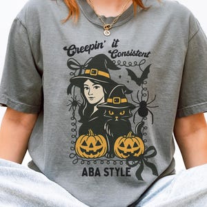 Puede incluir: Camiseta gris con un diseño de Halloween. El diseño presenta una bruja, un gato negro, calabazas, una araña y un murciélago. El texto "Creepin' it Consistent ABA STYLE" está impreso en la camiseta.