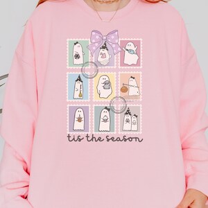 Puede incluir: Sudadera rosa claro con un diseño de ilustraciones de fantasmas dentro de marcos de sellos postales. Los sellos están dispuestos en forma de cuadrícula, con un lazo morado en la parte superior. El texto "tis the season" está impreso en la parte inferior.