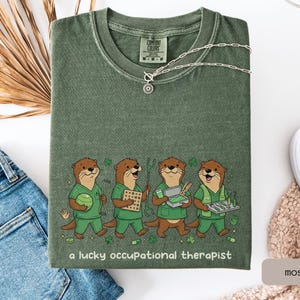 以下が含まれることがあります： モスグリーンのTシャツ。4匹のオッターが緑色のスクラブを着て、それぞれ作業療法ツールを持っています。「a lucky occupational therapist」の文字がオッターの下にプリントされています。シルバーのネックレスと白い靴も写っています。