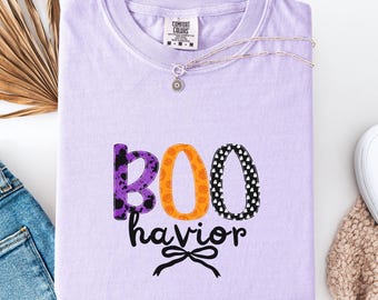 May include: A light purple t-shirt with the words "BOO havior" in a Halloween-themed design. The letters are in purple, orange, and black with patterns. A silver necklace and white sneakers are also visible.