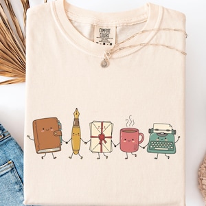 Op de afbeelding: Een crèmekleurig t-shirt met een afbeelding van vijf antropomorfe voorwerpen die elkaars hand vasthouden: een bruin dagboek, een gele vulpen, een witte envelop met een rood waszegel, een rode koffiemok en een groenblauwe typemachine.