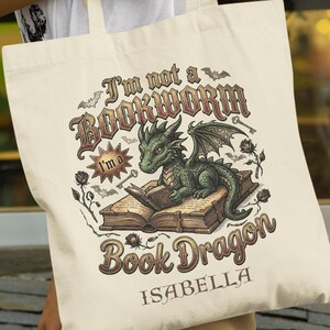 Puede incluir: Bolso tote color crema con un dragón verde leyendo un libro. El texto dice "I'm not a Bookworm, I'm a Book Dragon" con el nombre "Isabella" debajo. El diseño incluye murciélagos, llaves y rosas.