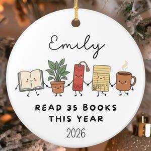 Puede incluir: Un adorno redondo de cerámica blanca con el nombre Emily en la parte superior, seguido de una fila de cinco simpáticos personajes de dibujos animados tomados de la mano: un libro, una planta en maceta, un marcapáginas, una ficha de biblioteca y una taza de café.