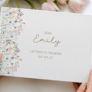 May include: A white hardcover book titled Dear Emily Letters to the Bride with the date 08.09.27 printed in gold. The left side of the cover features a vertical botanical illustration of small blue, yellow, and pink wildflowers.