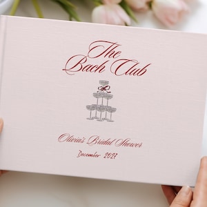 May include: A light pink hardcover book with the text The Bach Club in dark red script. Below the text is a grey illustration of a stacked champagne glass tower with a red bow. The text Olivia's Bridal Shower and December 2027 appear below.