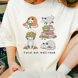 May include: Cream-colored t-shirt featuring a graphic of opossums in various reading poses. The opossums are depicted with books, blankets, and mugs. The text "Feral but well-read" is printed below the illustrations.