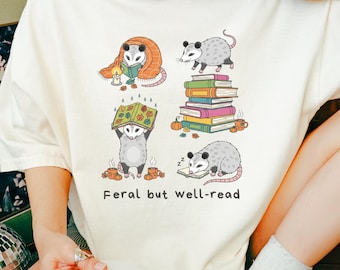 May include: Cream-colored t-shirt featuring a graphic of opossums in various reading poses. The opossums are depicted with books, blankets, and mugs. The text "Feral but well-read" is printed below the illustrations.