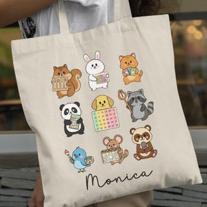 Puede incluir: Bolso tote beige con animales de dibujos animados, incluyendo una ardilla, un conejo, un gato, un panda, un perro, un mapache, un pájaro, un ratón y un oso. Cada animal sostiene un calendario o materiales de arte. El nombre "Monica" está escrito en cursiva en la parte inferior.