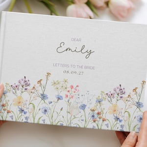 May include: A rectangular hardcover book with a light gray fabric cover featuring a colorful watercolor wildflower print along the bottom edge. The text reads DEAR Emily LETTERS TO THE BRIDE 08.09.27 in black and gray lettering on the front cover.