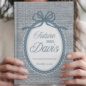 Puede incluir: Un cuaderno de tapa dura con textura de lino gris que presenta un marco ovalado blanco con un borde festoneado azul y un lazo azul. El texto dice Future MRS. Davis, LITTLE NOTES FOR THE BIG DAY y December 17, 2026 en color azul.
