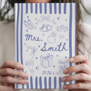 Puede incluir: Un cuaderno blanco con rayas verticales azules que presenta ilustraciones de línea azul de iconos de boda, incluyendo tacones, corazones, un anillo, un regalo y flores. El texto dice Mrs. Smith y el año 2026 en letra cursiva azul.