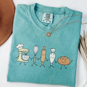 Puede incluir: Una camiseta de manga corta color verde azulado con un gráfico de una batidora, un batidor, un rodillo, una manga pastelera y una hogaza de pan sonrientes tomados de la mano. La camiseta tiene una etiqueta Comfort Colors en el cuello.