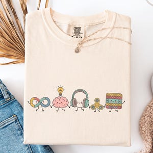 Comfort Colors Autism Doodle Characters Shirt featuring colorful, hand-drawn inclusive characters. Neatly styled next to classroom supplies, making it an ideal inclusion shirt, autism teacher shirt, and back to school shirt.