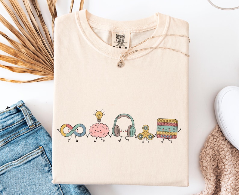 Comfort Colors Autism Doodle Characters Shirt featuring colorful, hand-drawn inclusive characters. Neatly styled next to classroom supplies, making it an ideal inclusion shirt, autism teacher shirt, and back to school shirt.