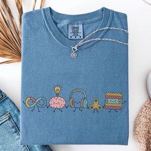 May include: A blue short-sleeve t-shirt with a Comfort Colors label at the neck. The front features a graphic of five cartoon characters holding hands: an infinity symbol, a brain with a lightbulb, headphones, a fidget spinner, and a square pop-it.