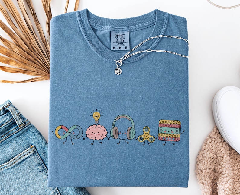 May include: A blue short-sleeve t-shirt with a Comfort Colors label at the neck. The front features a graphic of five cartoon characters holding hands: an infinity symbol, a brain with a lightbulb, headphones, a fidget spinner, and a square pop-it.