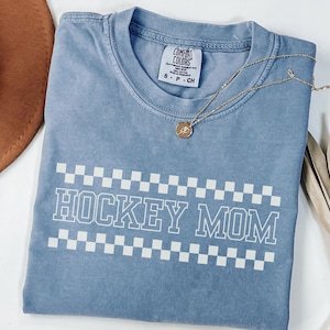 Puede incluir: Una camiseta azul Comfort Colors con las palabras "HOCKEY MOM" en letras blancas, enmarcada por un patrón de cuadros blancos y negros. Un collar dorado con un colgante descansa sobre la camisa. Un sombrero marrón y una planta seca están en el fondo.