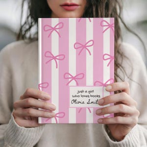 Puede incluir: Un cuaderno a rayas rosas y blancas con lazos rosas. El cuaderno tiene el texto "just a girl who loves books" y el nombre "Olivia Smit".