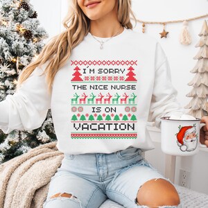 May include: White sweatshirt with a festive design. The text reads "I'M SORRY THE NICE NURSE IS ON VACATION" in red and green. A mug with a Santa Claus graphic is held in the hand.