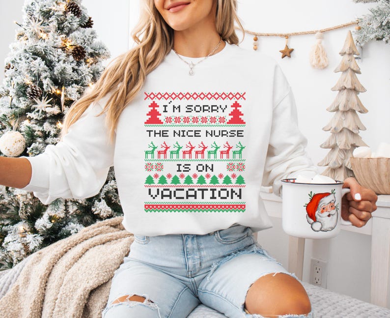 May include: White sweatshirt with a festive design. The text reads "I'M SORRY THE NICE NURSE IS ON VACATION" in red and green. A mug with a Santa Claus graphic is held in the hand.