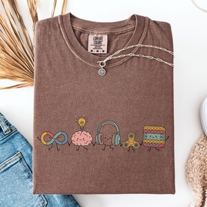 Comfort Colors Autism Doodle Characters Shirt featuring colorful, hand-drawn inclusive characters. Neatly styled next to classroom supplies, making it an ideal inclusion shirt, autism teacher shirt, and back to school shirt.