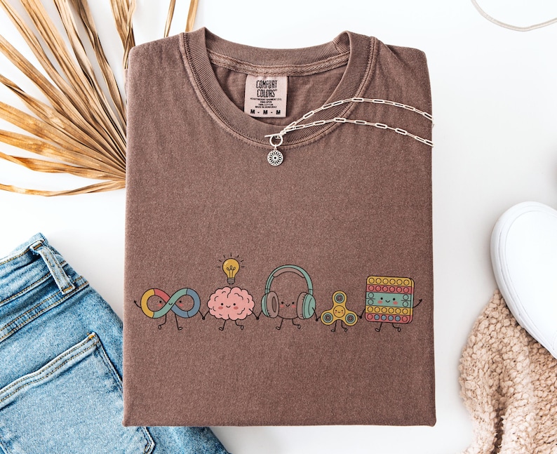 Comfort Colors Autism Doodle Characters Shirt featuring colorful, hand-drawn inclusive characters. Neatly styled next to classroom supplies, making it an ideal inclusion shirt, autism teacher shirt, and back to school shirt.