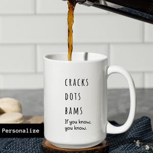 May include: White ceramic mug with the words "CRACKS DOTS BAMS" and the phrase "If you know, you know." Coffee is being poured into the mug. The mug is on a wooden coaster. The word "Personalize" is in a black box.