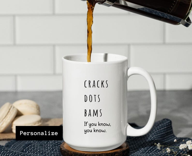 May include: White ceramic mug with the words "CRACKS DOTS BAMS" and the phrase "If you know, you know." Coffee is being poured into the mug. The mug is on a wooden coaster. The word "Personalize" is in a black box.