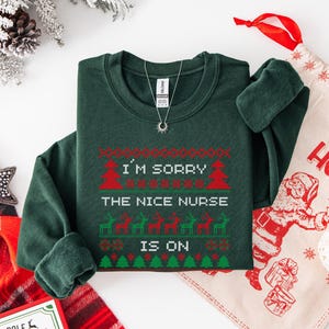 May include: A dark green sweatshirt with a festive design. The text "I'M SORRY THE NICE NURSE IS ON" is displayed in red and white, with Christmas tree and reindeer graphics. A silver necklace with a sun pendant is also visible.