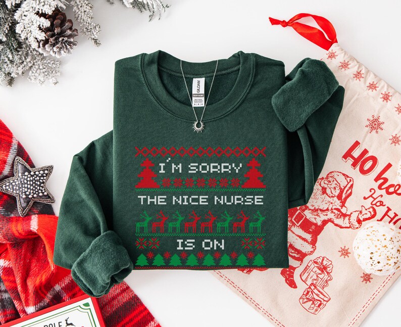 May include: A dark green sweatshirt with a festive design. The text "I'M SORRY THE NICE NURSE IS ON" is displayed in red and white, with Christmas tree and reindeer graphics. A silver necklace with a sun pendant is also visible.