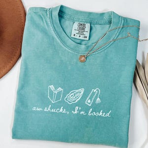 Puede incluir: Una camiseta de manga corta color verde azulado con un gráfico blanco que muestra un libro abierto, una ostra y un marcapáginas. El texto dice "aw shucks, I'm booked". La camiseta tiene una etiqueta Comfort Colors y un collar dorado.