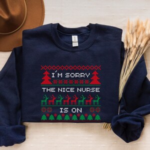 May include: Navy blue sweatshirt with a festive design. The text "I'M SORRY THE NICE NURSE IS ON" is displayed in a Christmas sweater style, with red, green, and white accents. The design includes Christmas trees and reindeer.
