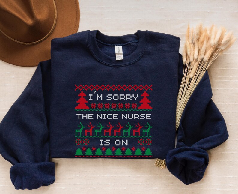 May include: Navy blue sweatshirt with a festive design. The text "I'M SORRY THE NICE NURSE IS ON" is displayed in a Christmas sweater style, with red, green, and white accents. The design includes Christmas trees and reindeer.