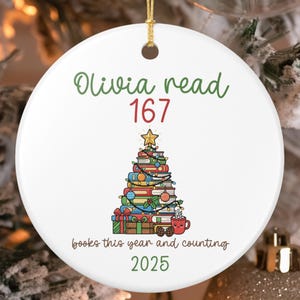 Puede incluir: Adorno navideño de cerámica blanca con una cuerda dorada. El adorno presenta el texto "Olivia read 167" en verde y rojo. Un árbol de Navidad hecho de libros está en el centro, con el texto "books this year and counting 2025" debajo.