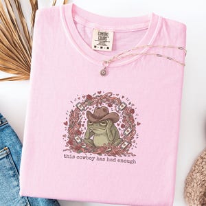 Puede incluir: Camiseta rosa Comfort Colors con un dibujo de una rana con sombrero de vaquero, rodeada de una corona floral con corazones y naipes. El texto "this cowboy has had enough" está debajo. Un collar dorado está sobre la camiseta.