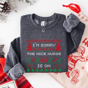 May include: Dark gray sweatshirt with a festive Christmas design. The text reads "I'M SORRY THE NICE NURSE IS ON" in red and green. The design includes Christmas trees and reindeer. A silver necklace with a sun pendant is visible.