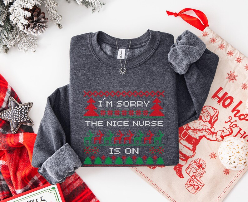 May include: Dark gray sweatshirt with a festive Christmas design. The text reads "I'M SORRY THE NICE NURSE IS ON" in red and green. The design includes Christmas trees and reindeer. A silver necklace with a sun pendant is visible.