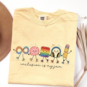 Puede incluir: Una camiseta de color amarillo pálido con un gráfico de cinco personajes de dibujos animados tomados de la mano: un símbolo de infinito arcoíris, un cerebro rosa, un juguete pop-it arcoíris, auriculares azules y un lápiz amarillo. Texto "inclusion is my jam".