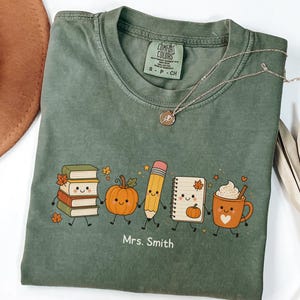 Puede incluir: Una camiseta de manga corta color verde oliva con un gráfico de una pila de libros, una calabaza, un lápiz, un cuaderno de espiral y una taza de café, todos con caras sonrientes. El texto "Mrs. Smith" está impreso debajo en letras blancas.