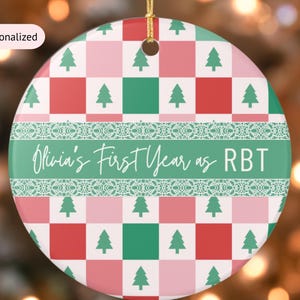Puede incluir: Adorno navideño redondo de cerámica con un patrón a cuadros rojo, verde y rosa y árboles de Navidad verdes. El adorno tiene una banda verde con el texto "Olivia's First Year as RBT" en escritura blanca. Se adjunta una cuerda dorada.