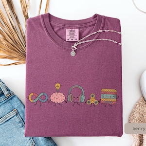 May include: A berry-colored t-shirt featuring a graphic of five cute, anthropomorphic characters holding hands: an infinity symbol, a brain with a lightbulb, headphones, a fidget spinner, and a square pop-it toy. The shirt has a Comfort Colors label.