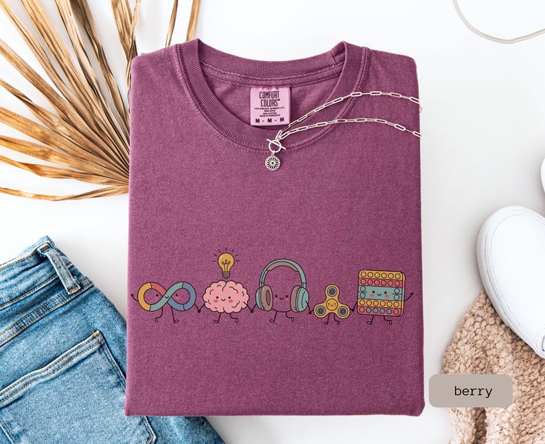 May include: A berry-colored t-shirt featuring a graphic of five cute, anthropomorphic characters holding hands: an infinity symbol, a brain with a lightbulb, headphones, a fidget spinner, and a square pop-it toy. The shirt has a Comfort Colors label.