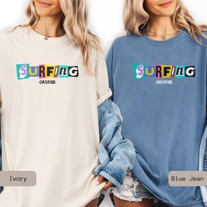 May include: Two t-shirts, one ivory and one blue jean, featuring the word "SURFING" in colorful, blocky letters. Below the word is the name "JACKIE". The shirts are casual, with rolled-up sleeves.