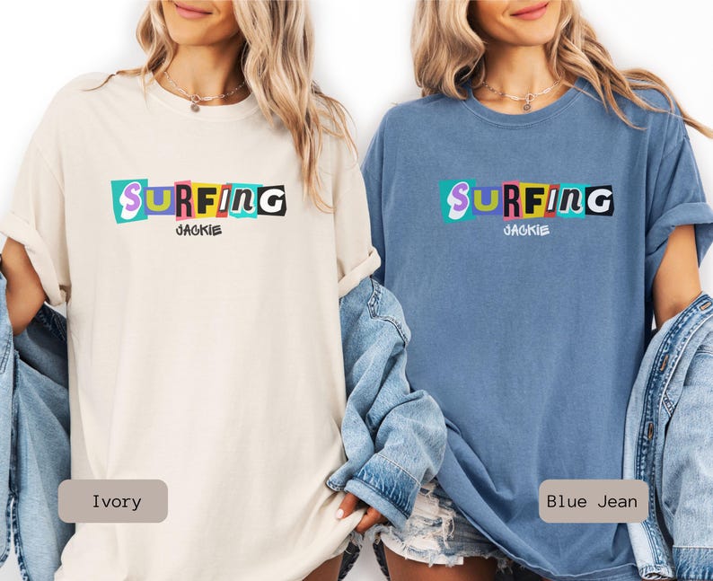 May include: Two t-shirts, one ivory and one blue jean, featuring the word "SURFING" in colorful, blocky letters. Below the word is the name "JACKIE". The shirts are casual, with rolled-up sleeves.